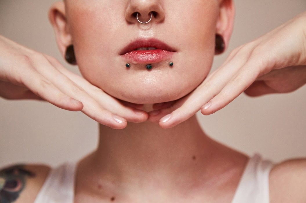 Woman with piercings and a neutral expression, red lips, hands on chin, white clothing.