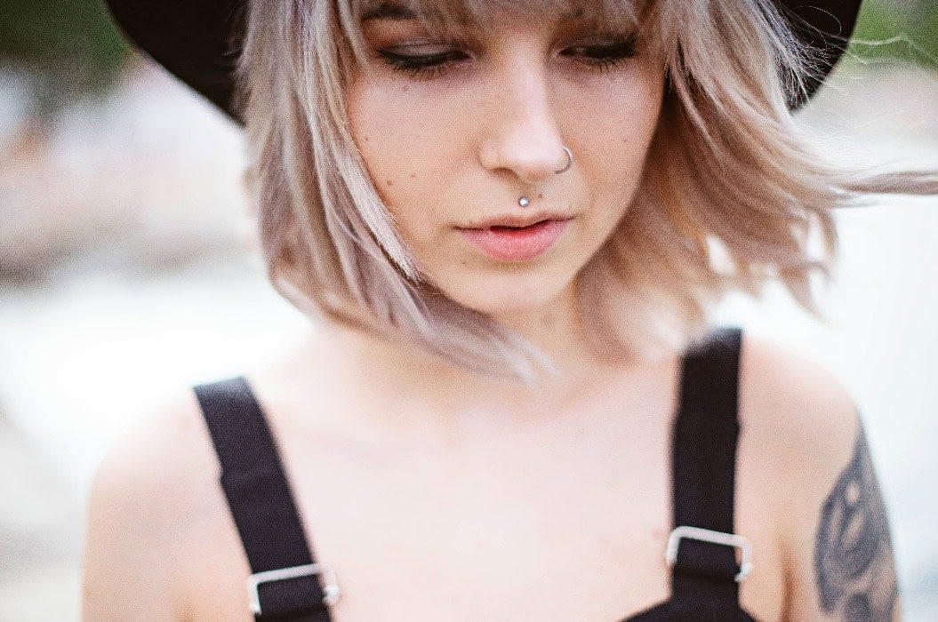 Junge Frau mit nachdenklichem Blick, blonden Haaren, schwarzem Hut und Nasenpiercing.