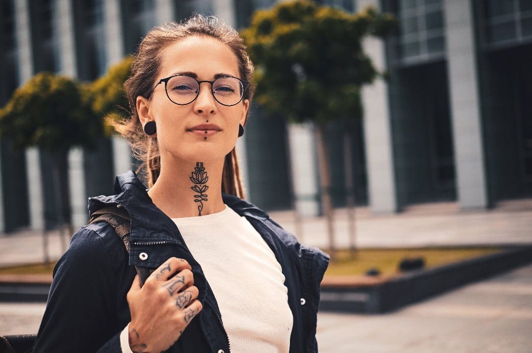 Frau mit Brille, neutralem Gesichtsausdruck, Tattoos, hellem Shirt, dunkler Jacke.